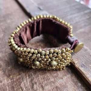 Peut inclure: Un bracelet de perles de couleur or avec un intérieur marron. Le bracelet comporte de petites perles rondes et de plus grosses perles décoratives. Le bracelet est sur une surface en bois.