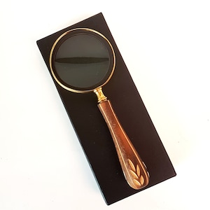 May include: A gold magnifying glass with a wooden handle featuring a carved leaf design. The magnifying glass is on a black background.
