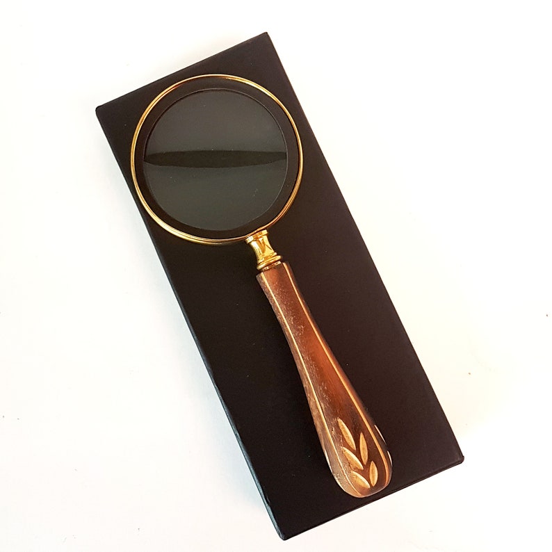 May include: A gold magnifying glass with a wooden handle featuring a carved leaf design. The magnifying glass is on a black background.