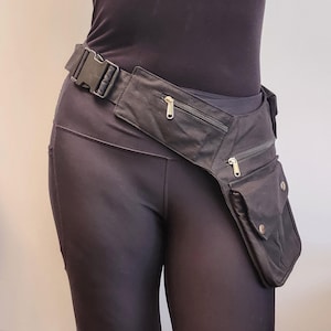 Peut inclure: Une ceinture utilitaire noire dotée de plusieurs compartiments de rangement, dont deux poches zippées et une poche à rabat avec fermetures à pression. La ceinture est fixée par une boucle latérale en plastique noir et se porte à la taille.