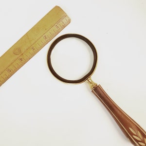 May include: A magnifying glass with a gold-coloured frame and a wooden handle with a floral design. The handle is brown with a light brown floral pattern.