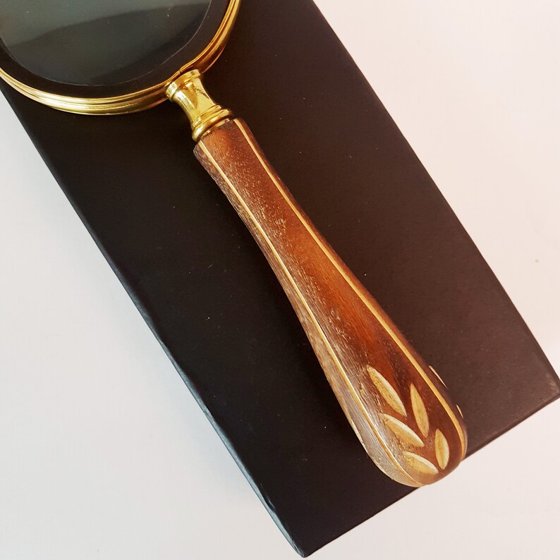 May include: A magnifying glass with a brass frame and a wooden handle with a carved leaf design. The handle is a dark brown colour and the brass frame is a shiny gold colour.