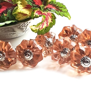 May include: A set of eight pink glass cabinet knobs with a flower design and silver accents. The knobs are arranged in a row on a white surface.