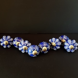 May include: Set of eight blue ceramic knobs with white floral designs and gold accents. The knobs are arranged in a semi-circle on a black surface.