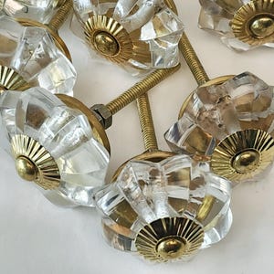 May include: A set of clear glass cabinet knobs with gold metal accents. The knobs have a faceted design and are attached to a screw.
