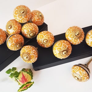 May include: A collection of yellow ceramic cabinet knobs with white floral designs. The knobs have a gold metal accent in the centre.