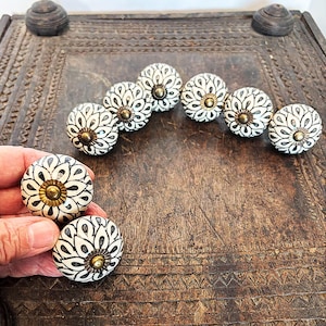 May include: A collection of round ceramic knobs featuring a black and white petal pattern radiating from a central brass-toned metal cap. The knobs have a smooth, rounded surface and are arranged in a semi-circular formation on a dark surface.