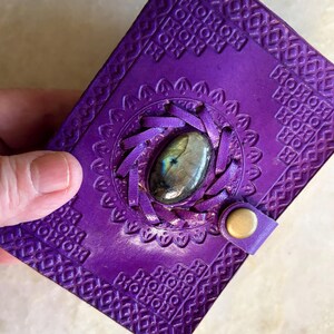 Puede incluir: Un diario rectangular de cuero morado con un patrón geométrico en relieve en la cubierta. Un recorte circular central muestra una piedra preciosa de labradorita ovalada rodeada por tiras de cuero trenzado. Un pequeño cierre de latón asegura la cubierta.