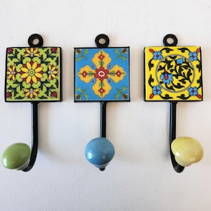 May include: Three decorative wall hooks with colourful ceramic tiles. The tiles feature floral and geometric patterns in shades of blue, green, yellow, and red. The hooks are black metal with a rounded knob at the bottom.
