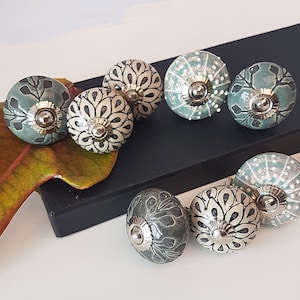 May include: Assortment of ceramic cabinet knobs with a variety of patterns in shades of green and white. The knobs have a silver metal accent in the center.
