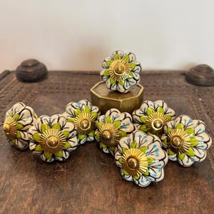 May include: A collection of nine ceramic drawer knobs with a scalloped, flower-like shape. Each knob features a white base with hand-painted lime green and black leaf patterns, blue accents, and a central circular gold-toned metal hardware piece.