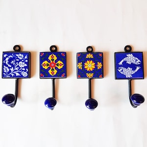 Könnte beinhalten: Vier blaue und gelbe Keramik-Haken mit schwarzen Metallhaltern. Jede Fliese zeigt ein anderes Design, darunter ein Blumenmuster, ein Kreuz, eine Blume und zwei Fische.