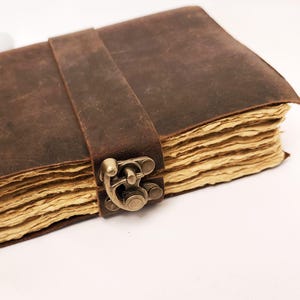 May include: A brown leather journal with a strap closure and antique brass clasp. The pages are aged and have a textured appearance. The journal is closed and ready to be written in.