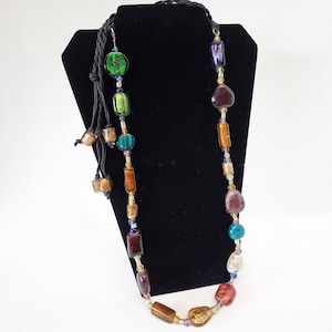 May include: A long multi-colored glass bead necklace with a black cord and tassels. The beads are various shapes and sizes, including round, square, and oval. The colors include green, blue, purple, brown, and gold.