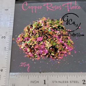 May include: A close-up of a pile of iridescent copper rose flake glitter, weighing 0.25 ounces, against a grey background. The glitter is a mix of green, gold, and pink.