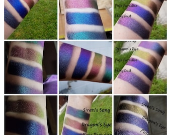 May include: A close-up of four different eyeshadows swatched on a person's arm. The eyeshadows are labeled "Siren's Song", "Dragon's Eye", "Pixie Paradise", and "Fairy Dust".