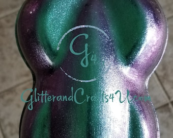 Op de afbeelding: Een close-up van groene, paarse en zilveren iriserende glitter. De glitter zit in een kleine, ronde container. De container heeft een witte cirkel met de letter "G" en het nummer "4" erin. De tekst "GlitterandCrafts4U.com" is zichtbaar onder de cirkel.