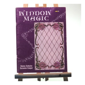 May include: A purple book titled "Window Magic" with a stained glass design featuring roses and a diamond pattern. The book is by Donna Schulze and Suzanne Cooper.