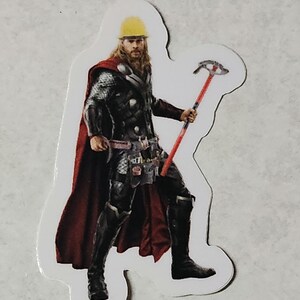 Pode incluir: Um adesivo com um super-her&oacute;i usando um capacete amarelo e uma capa vermelha, segurando uma ferramenta vermelha e prateada.