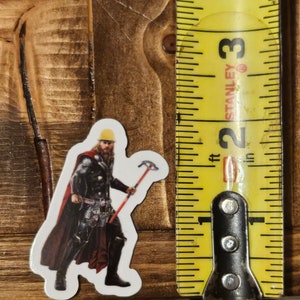 Pode incluir: Um adesivo de vinil de um super-her&oacute;i de desenho animado usando um capacete amarelo, uma capa vermelha e segurando um martelo. O adesivo est&aacute; em uma superf&iacute;cie de madeira ao lado de uma fita m&eacute;trica.