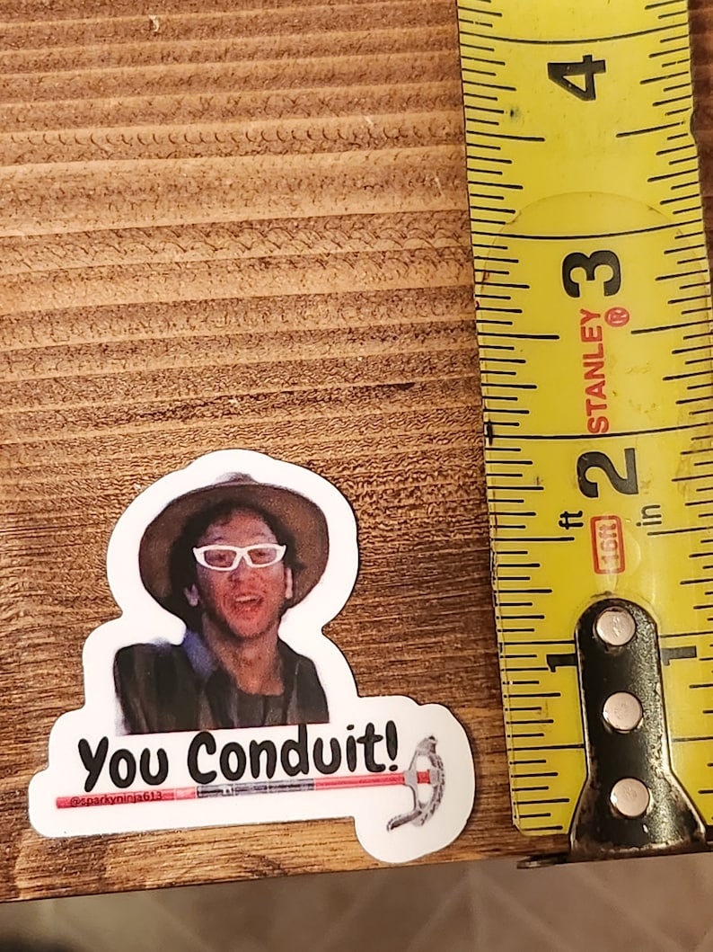 K&ouml;nnte beinhalten: Ein wei&szlig;er Aufkleber mit einer Cartoon-Abbildung einer Person, die einen Hut, eine Sonnenbrille und eine Jacke tr&auml;gt. Der Aufkleber tr&auml;gt den Text "You Conduit!" und eine kleine rote und wei&szlig;e Hammergrafik. Der Aufkleber befindet sich auf einer Holzoberfl&auml;che.