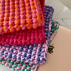 May include: A stack of four crocheted squares in different colors, including pink, orange, purple, and green. The squares are stacked on top of a pink laptop.
