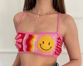 Puede incluir: Un top de crochet rosa con un smiley, una flor y un arcoíris. El top tiene tirantes finos y es corto.