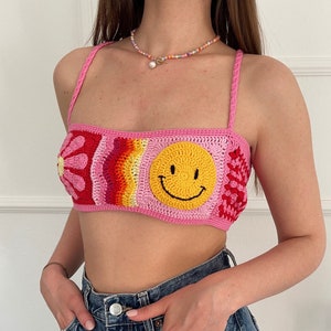 以下が含まれることがあります： スマイリー、花、虹のデザインが施されたピンクのクロシェトップ。トップは細いストラップで、クロップド丈です。