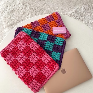 Może przedstawiać: Trzy kolorowe, szydełkowe etui na laptopa w odcieniach różu, czerwieni, pomarańczy, fioletu i turkusu. Etui mają wzór w szachownicę i są prezentowane na białym stole obok złotego laptopa. Marka "MARCELLINE" jest widoczna na metce.