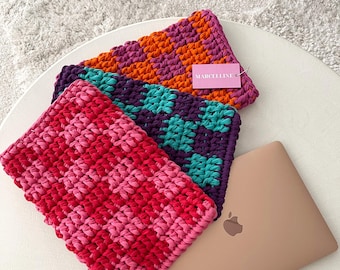 May include: Three colorful, crocheted laptop sleeves in shades of pink, red, orange, purple, and teal. The sleeves have a checkered pattern and are displayed on a white table next to a gold laptop. The brand name "MARCELLINE" is visible on a tag.