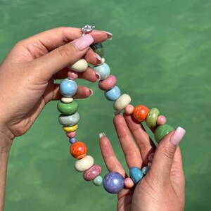 Puede incluir: Un collar de cuentas de cer&aacute;mica de colores con una variedad de colores, incluyendo verde, azul, rosa, naranja y amarillo. Las cuentas son de diferentes tama&ntilde;os y formas, y est&aacute;n enhebradas en una cadena de plata.