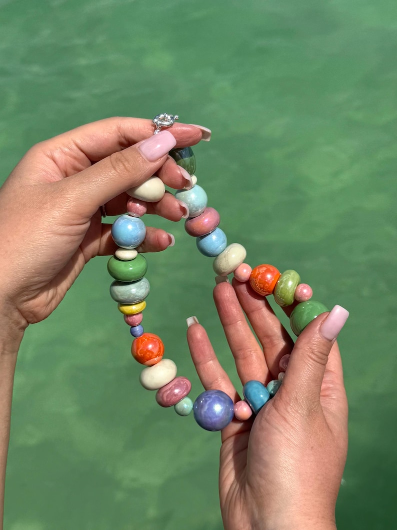 Puede incluir: Un collar de cuentas de cer&aacute;mica de colores con una variedad de colores, incluyendo verde, azul, rosa, naranja y amarillo. Las cuentas son de diferentes tama&ntilde;os y formas, y est&aacute;n enhebradas en una cadena de plata.