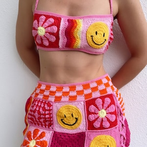 May include: A pink crochet skirt with a patchwork design featuring smiley faces, flowers, and checkered patterns.