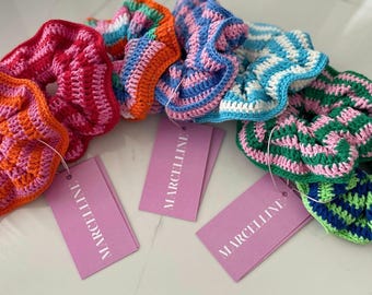 May include: A set of colorful crocheted hair scrunchies with pink, blue, green, and orange stripes. The scrunchies are arranged on a white surface with pink tags that say "MARCELLINE".