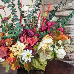 May include: A fall-themed floral arrangement in a green vase. The arrangement features red, white, and yellow flowers, along with green leaves and red berries.