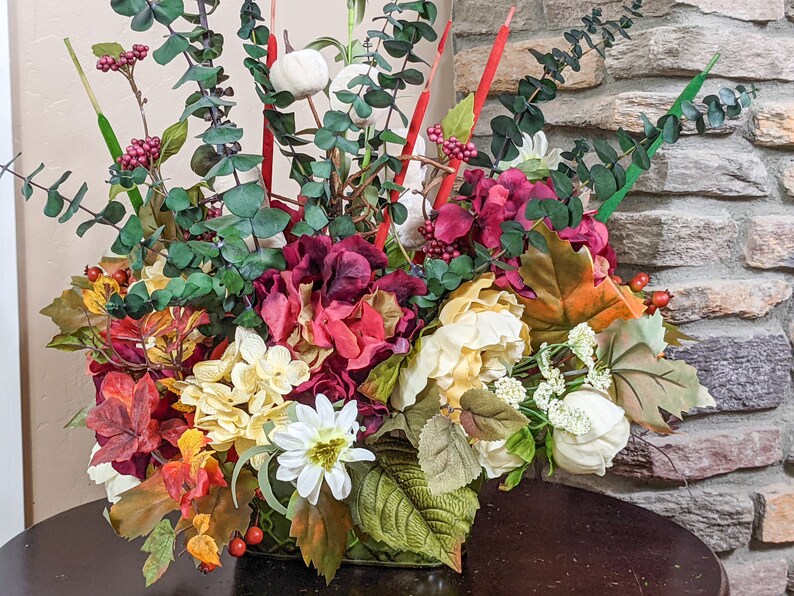 May include: A fall-themed floral arrangement in a green vase. The arrangement features red, white, and yellow flowers, along with green leaves and red berries.