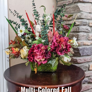 May include: Multi-colored fall centerpiece with red, white, and green flowers in a green woven basket. The centerpiece is on a dark brown wooden table.