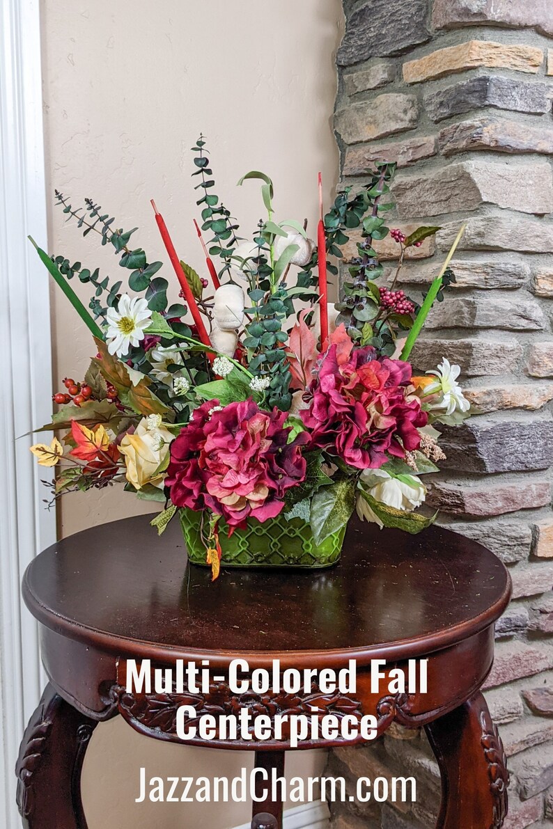 May include: Multi-colored fall centerpiece with red, white, and green flowers in a green woven basket. The centerpiece is on a dark brown wooden table.