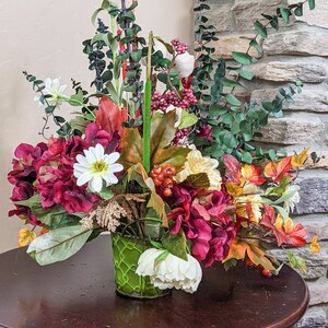 May include: A fall-themed floral arrangement in a green wire basket. The arrangement features burgundy hydrangeas, white daisies, yellow flowers, green eucalyptus, red berries, and fall leaves.