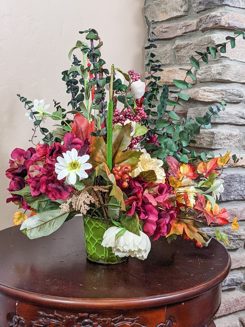 May include: A fall-themed floral arrangement in a green wire basket. The arrangement features burgundy hydrangeas, white daisies, yellow flowers, green eucalyptus, red berries, and fall leaves.