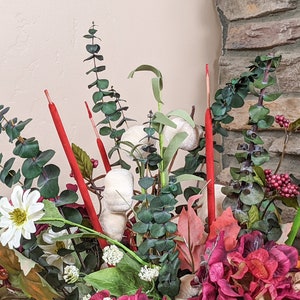 May include: A floral arrangement in a vase with red, white, and green stems, white cotton balls, and red and green berries. The arrangement is in front of a stone wall.