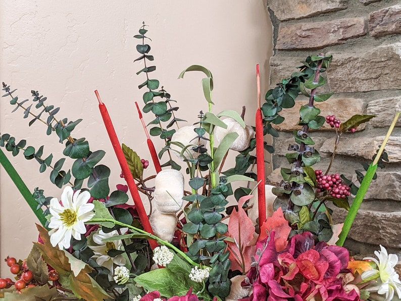 May include: A floral arrangement in a vase with red, white, and green stems, white cotton balls, and red and green berries. The arrangement is in front of a stone wall.