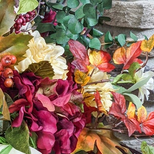May include: A bouquet of artificial flowers with burgundy, white, and yellow blooms. The bouquet also includes green leaves, red berries, and orange and brown fall leaves.
