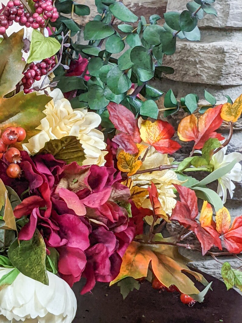 May include: A bouquet of artificial flowers with burgundy, white, and yellow blooms. The bouquet also includes green leaves, red berries, and orange and brown fall leaves.