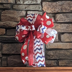 May include: A red, white and blue bow with a chevron pattern and white polka dots. The bow is tied with a red ribbon and has a white star on each side.