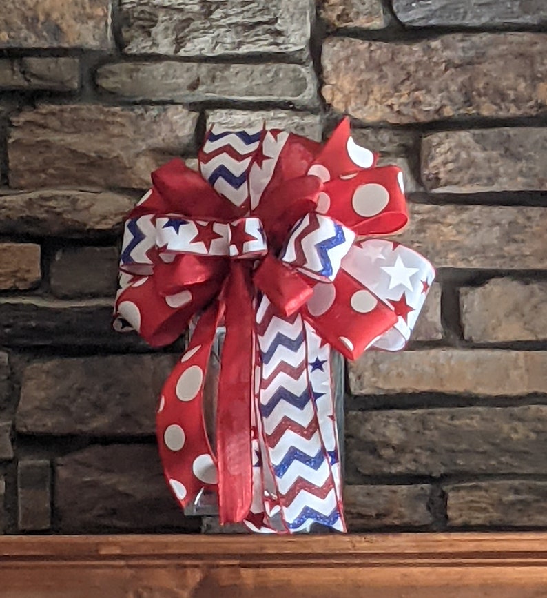 May include: A red, white and blue bow with a chevron pattern and white polka dots. The bow is tied with a red ribbon and has a white star on each side.