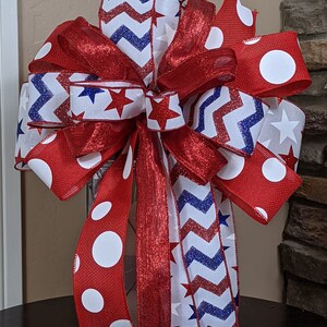 May include: A red, white and blue bow with a chevron pattern, white stars and white polka dots. The bow is tied around a clear glass lantern.