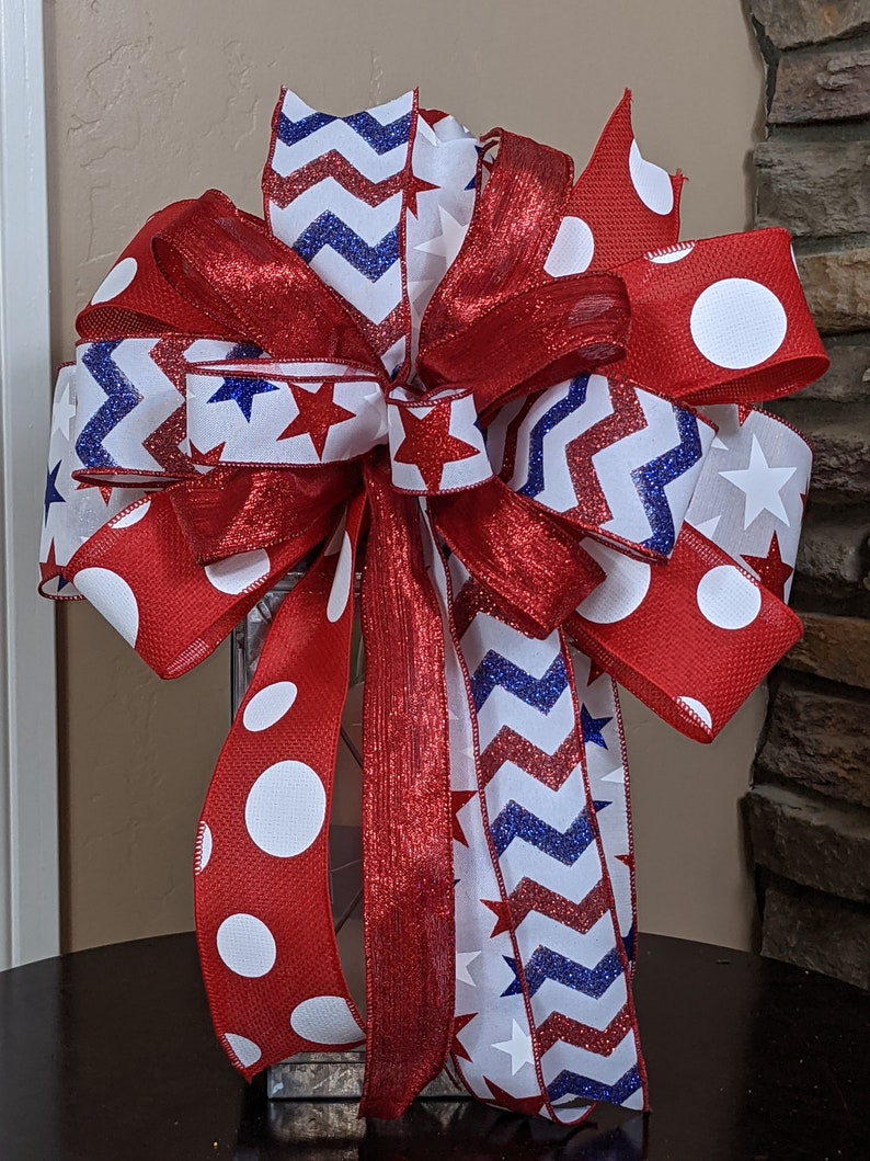 May include: A red, white and blue bow with a chevron pattern, white stars and white polka dots. The bow is tied around a clear glass lantern.