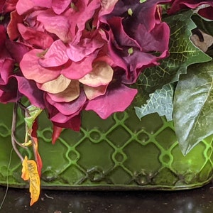 May include: A green planter with a lattice design, filled with a bouquet of pink and purple flowers and green leaves.