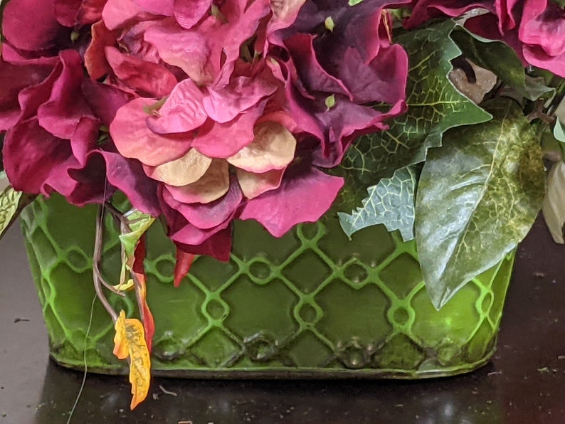May include: A green planter with a lattice design, filled with a bouquet of pink and purple flowers and green leaves.